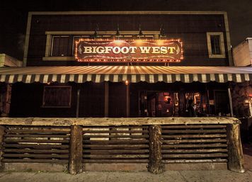 Nighttime exterior of a rustic western saloon with a glowing marquee sign, striped awning, log rail fence and warm doorway lighting