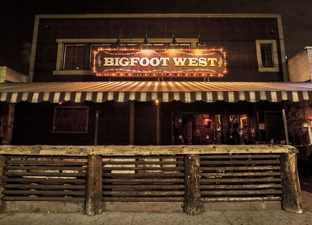 Nighttime exterior of a rustic western saloon with a glowing marquee sign, striped awning, log rail fence and warm doorway lighting