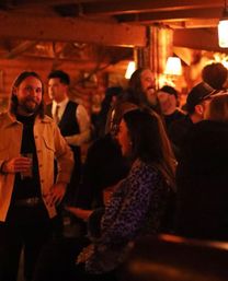Cozy, dimly lit rustic bar interior with wooden beams, people chatting and holding drinks under warm amber lighting — lively nightlife scene.