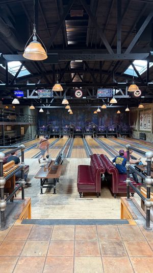 Retro industrial bowling alley interior with wooden lanes, leather seating, bowlers in action under exposed timber trusses, pendant lights and skylights