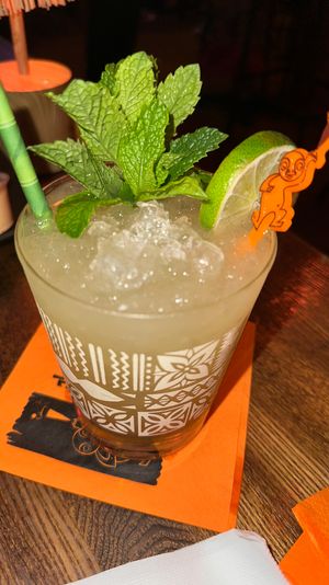 Tropical mint-lime cocktail in a patterned tiki glass with crushed ice, fresh mint sprig, lime wedge and a playful orange stirrer, served on an orange napkin on a wooden bar table.