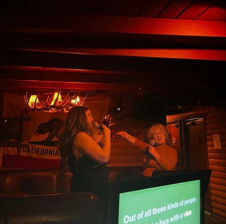 Two people singing a karaoke duet under an antler chandelier in a cozy, red-lit rustic bar with a California flag backdrop.