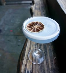 Foamy pale blue cocktail in a coupe glass topped with a dried citrus wheel, sitting on a weathered wooden railing outdoors
