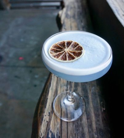 Foamy pale blue cocktail in a coupe glass topped with a dried citrus wheel, sitting on a weathered wooden railing outdoors