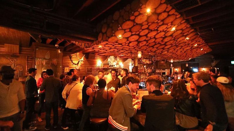 Busy rustic cocktail bar interior with round wood-slab ceiling panels, warm amber lighting, bartenders serving drinks and patrons socializing at a crowded bar