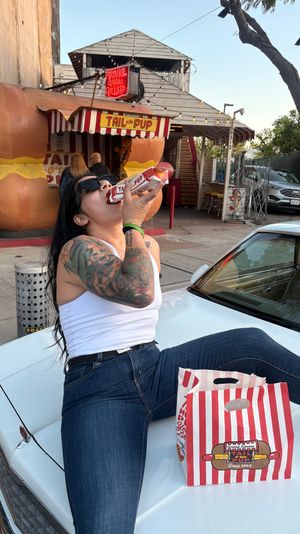 Person with sunglasses and colorful arm tattoo sitting on a car hood, eating a hot dog from a red-and-white striped takeout bag in front of a retro oversized-bun hot dog stand with a neon sign.