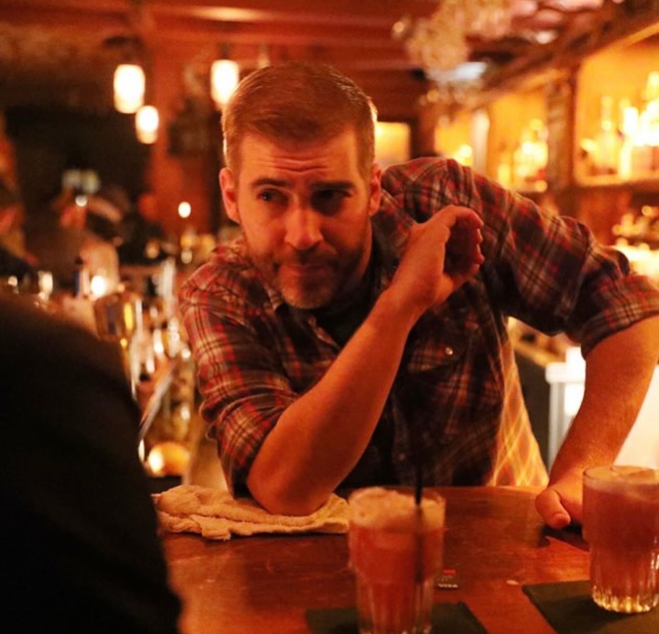 Person in a plaid shirt leaning on a wooden bar in a warm, dimly lit cozy pub with two cocktails in front.