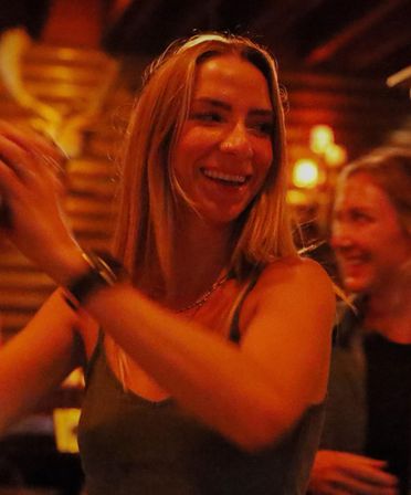 Blonde woman laughing and clapping during a night out in a dimly lit cozy bar with warm amber lighting and a smiling friend blurred in the background