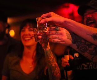 Friends clinking shot glasses in a dim red-lit bar — tattooed arms and smiling faces mid-toast, lively nightlife scene