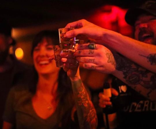 Friends clinking shot glasses in a dim red-lit bar — tattooed arms and smiling faces mid-toast, lively nightlife scene