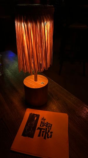 Glowing tiki-style table lamp with woven fringe casting warm orange light over a wooden table, a nearby glass and retro tiki-themed napkin in a dim bar setting.