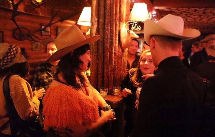 Cozy dimly lit log-cabin bar scene with patrons mingling and sipping drinks; two people in cowboy hats chat by a wooden post with a mounted deer head.