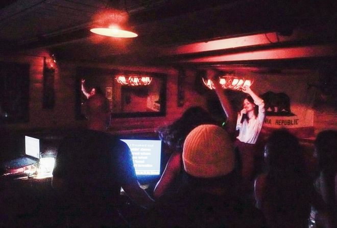 Intimate downtown karaoke night in a dimly lit bar, singer on a small stage under red pendant lights with raised hands from the crowd, laptop lyric screens and silhouetted patrons in the foreground.