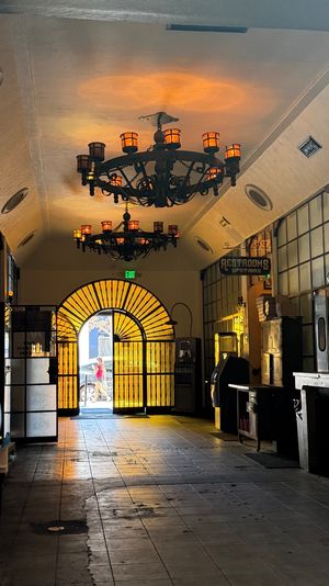 Historic lobby with tiled floor and wrought-iron chandeliers, golden light streaming through a yellow arched iron gate and a pedestrian silhouette outside.