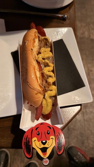 Gourmet grilled hot dog topped with mustard and caramelized onions in a toasted bun, served in a cardboard tray on a white plate at a restaurant table with a playful cartoon dog tag