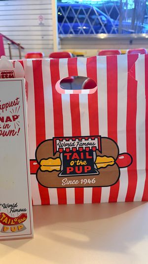 Retro red-and-white striped hot dog takeout bag with cartoon hot dog logo and matching tall white carton on a diner table, red diner chairs and window lights blurred in the background.