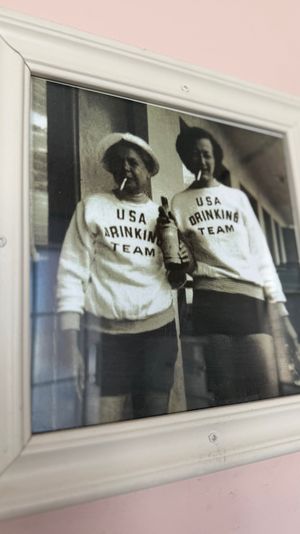 Framed vintage black-and-white photo of two people in matching 'USA Drinking Team' sweaters holding a bottle and cigarettes, displayed as wall art on a pale pink wall.