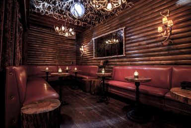 Candlelit cozy log-cabin lounge interior with red leather banquettes, round wood-stump side tables, antler chandeliers and dark wood-paneled walls.