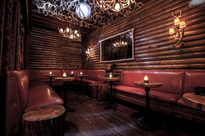 Candlelit cozy log-cabin lounge interior with red leather banquettes, round wood-stump side tables, antler chandeliers and dark wood-paneled walls.