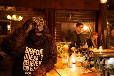 Costumed Bigfoot wearing a 'Bigfoot doesn't believe in you either' T-shirt at a cozy rustic log-cabin bar, leaning on a candlelit wooden counter with drinks and patrons in the background.