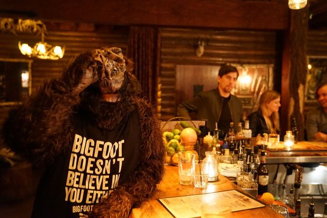 Someone in a Bigfoot costume wearing a "BIGFOOT DOESN'T BELIEVE IN YOU EITHER" shirt at a cozy rustic wooden bar with drinks, a fruit basket, and patrons in warm dim lighting.
