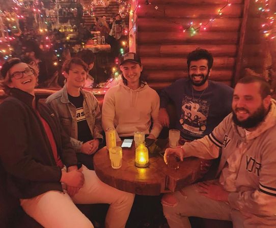 Five friends laughing around a small round wooden table in a cozy log-cabin style bar with colorful string lights and mirror reflections, drinks and a glowing candle jar on the table.