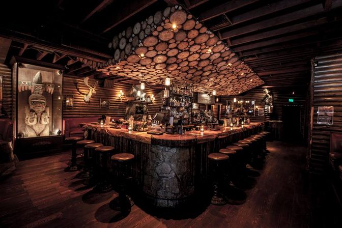 Cozy rustic log-cabin bar interior with a large central wooden-slab bar and round stools, log-slice ceiling feature, warm amber lighting, stacked bottles, mounted deer and carved totem—mountain lodge vibe.