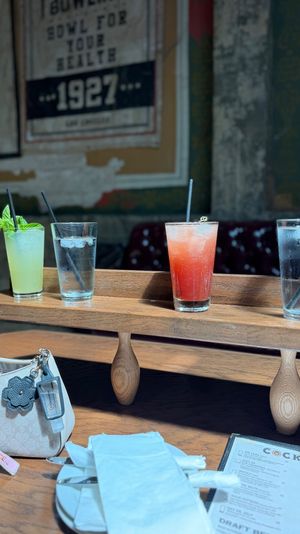 Sunlit cocktail flight on a wooden tasting board in a rustic bar: minty green drink, two waters and a red cocktail beside a purse and menu.