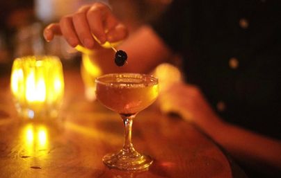 Candlelit bar scene featuring a coupe glass cocktail with a dark cherry on a pick, warm amber candlelight reflecting on the wooden bar.