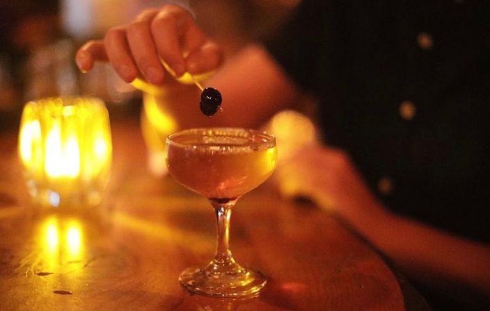 Candlelit bar scene featuring a coupe glass cocktail with a dark cherry on a pick, warm amber candlelight reflecting on the wooden bar.