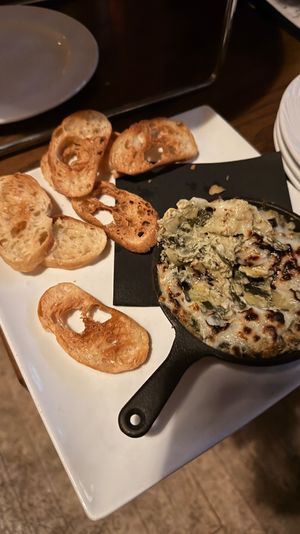 Warm spinach and artichoke dip with melted browned cheese in a small cast-iron skillet served with toasted baguette crostini on a white plate — restaurant appetizer