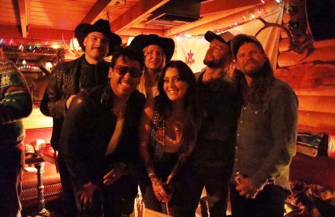 Group of seven friends smiling for a night-out photo inside a dim, wood-paneled rustic bar with cowboy hats, string lights and cozy western decor