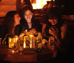 Three friends in a cozy candlelit bar booth, laughing at smartphones over a table scattered with beer cans and glasses — warm nightlife vibe.