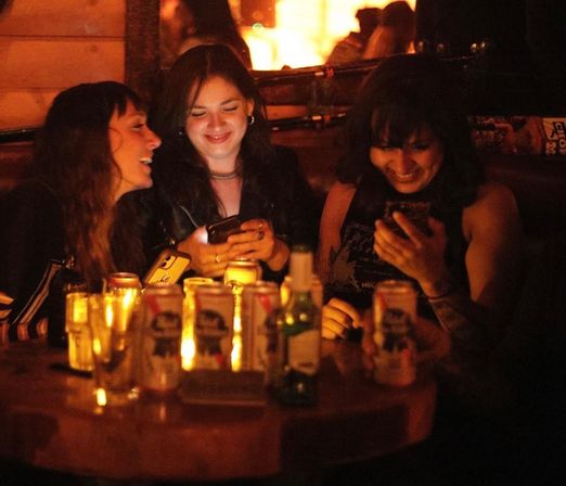 Three friends in a cozy candlelit bar booth, laughing at smartphones over a table scattered with beer cans and glasses — warm nightlife vibe.