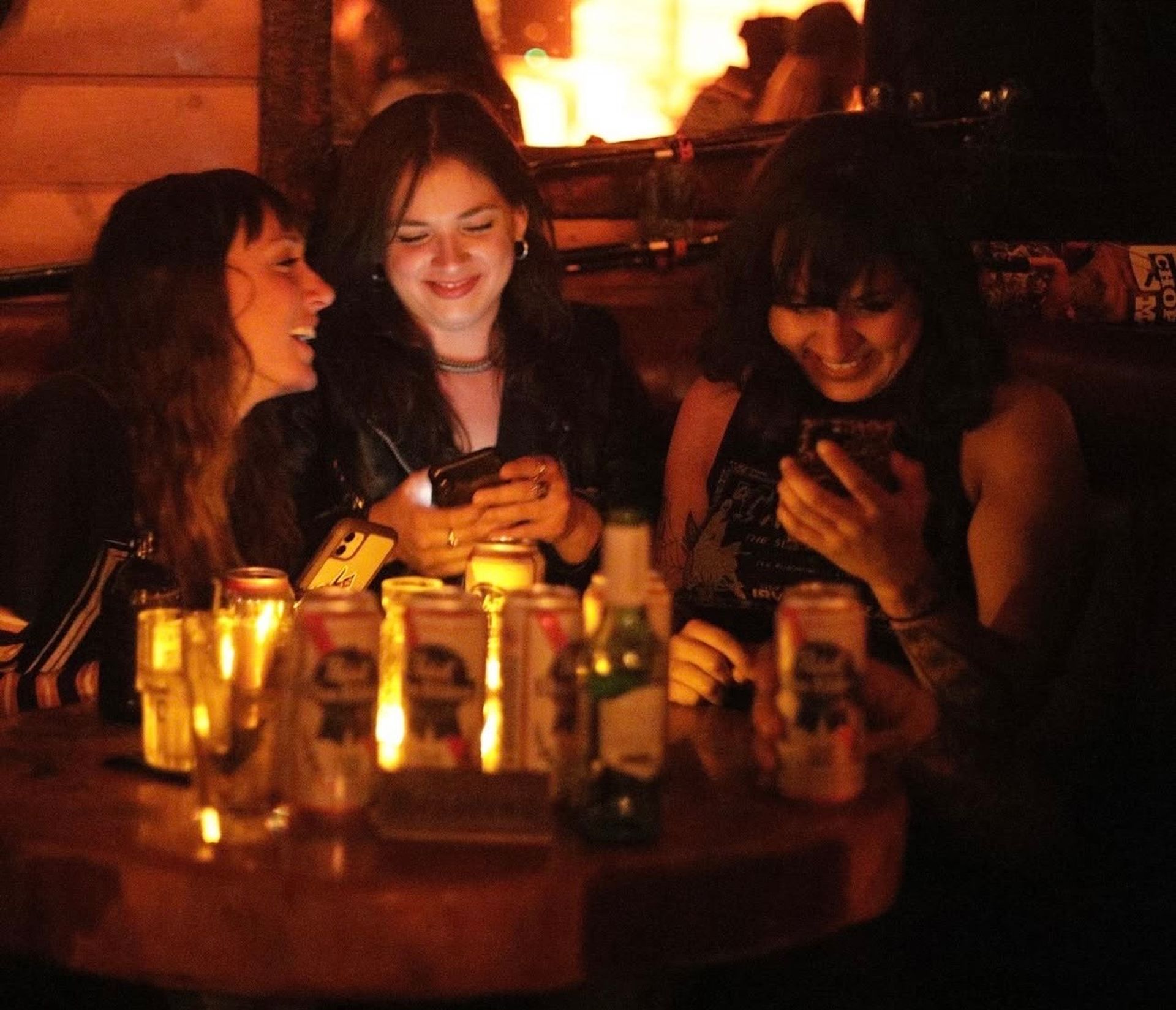 Three friends in a cozy candlelit bar booth, laughing at smartphones over a table scattered with beer cans and glasses — warm nightlife vibe.