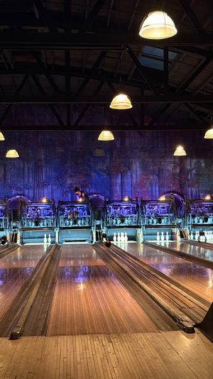 Vintage bowling alley interior with polished wooden lanes, antique mechanical pinsetters and rows of glowing pendant lights casting moody blue-purple ambient light over the pins