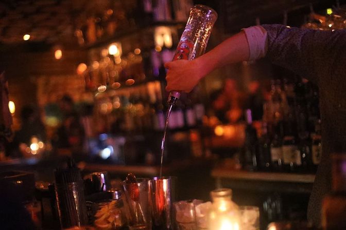 Bartender’s hand pouring liquor from a bottle into a metal cocktail shaker at a cozy, dimly lit bar with warm bokeh lights and a candlelit counter.