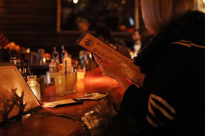 Patron browsing a cocktail menu at a cozy, dimly lit bar with lemon-garnished cocktails and bottles on a warm wooden counter.
