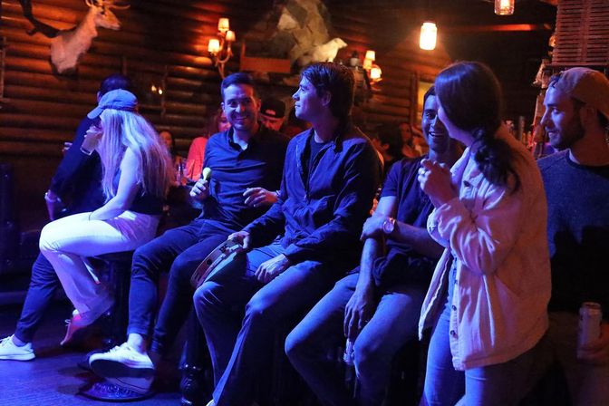 Nightlife scene in a rustic log‑cabin bar: a group of friends laughing at the bar under warm lamps and blue stage lights, playing hand percussion (tambourine and maracas) and holding drinks.
