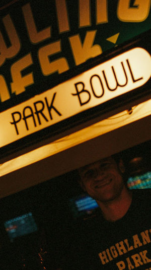 Warm-lit retro bowling alley marquee with glowing oval sign reading 'BOWL' above dim indoor lanes, neon colors and a smiling patron underneath.