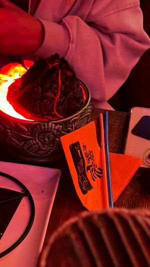 Cozy restaurant table close-up with a glowing red lava-rock centerpiece in a carved ceramic bowl, orange napkin and menu, metal straw, smartphone, and a pink-hooded sleeve nearby.