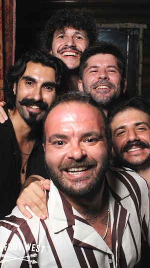Five men with beards and mustaches smiling closely in a lively indoor photo‑booth style portrait, one wearing a striped shirt in the foreground