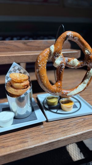 Giant salted soft pretzel on a metal stand next to crispy onion rings in a checkered-lined metal cup with three dipping sauces on white plates, served on a wooden pub table
