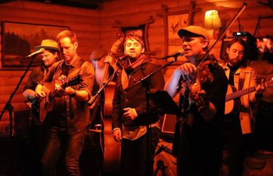 Five-member folk/bluegrass band playing acoustic guitars, upright bass, and fiddle on a small wood-paneled stage under warm orange lighting
