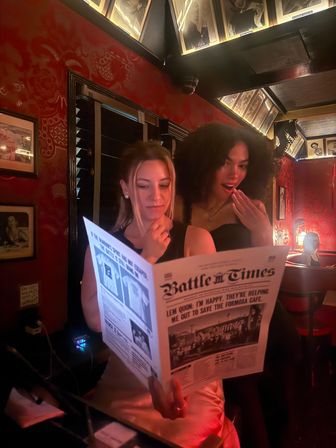 Two friends in a dim red-walled speakeasy-style bar reading a large broadsheet titled “Battle Times,” one looking surprised, with framed photos, warm low lighting and cozy booth seating — intimate nightlife scene.