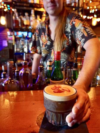 Bartender’s hand placing a frothy espresso-style cocktail topped with sliced strawberry garnish in a short glass on a wooden bar, colorful bottles and string lights blurred in the background.