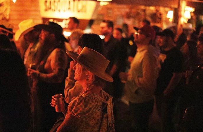 Crowd at a rustic saloon-style bar watching live music, foreground woman in a wide-brim hat holding a drink, warm amber lighting