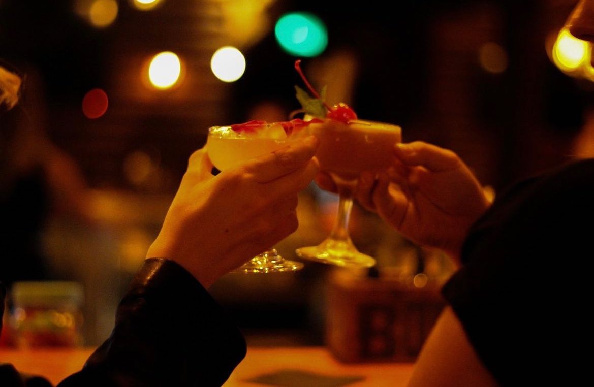 Two hands clinking garnished coupe cocktails in a warm, dimly lit bar with colorful bokeh lights — evening cocktail toast.