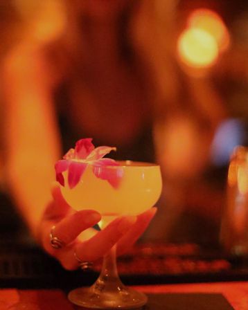 Hand holding a coupe glass with a pale yellow cocktail topped with pink orchid petals, warm orange bokeh bar ambiance