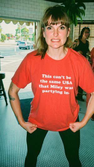 Candid photo of a woman in a red slogan T‑shirt making a playful grimace and flexing fists inside a bright café with hexagon tile floor and windows showing parked cars on a city street.